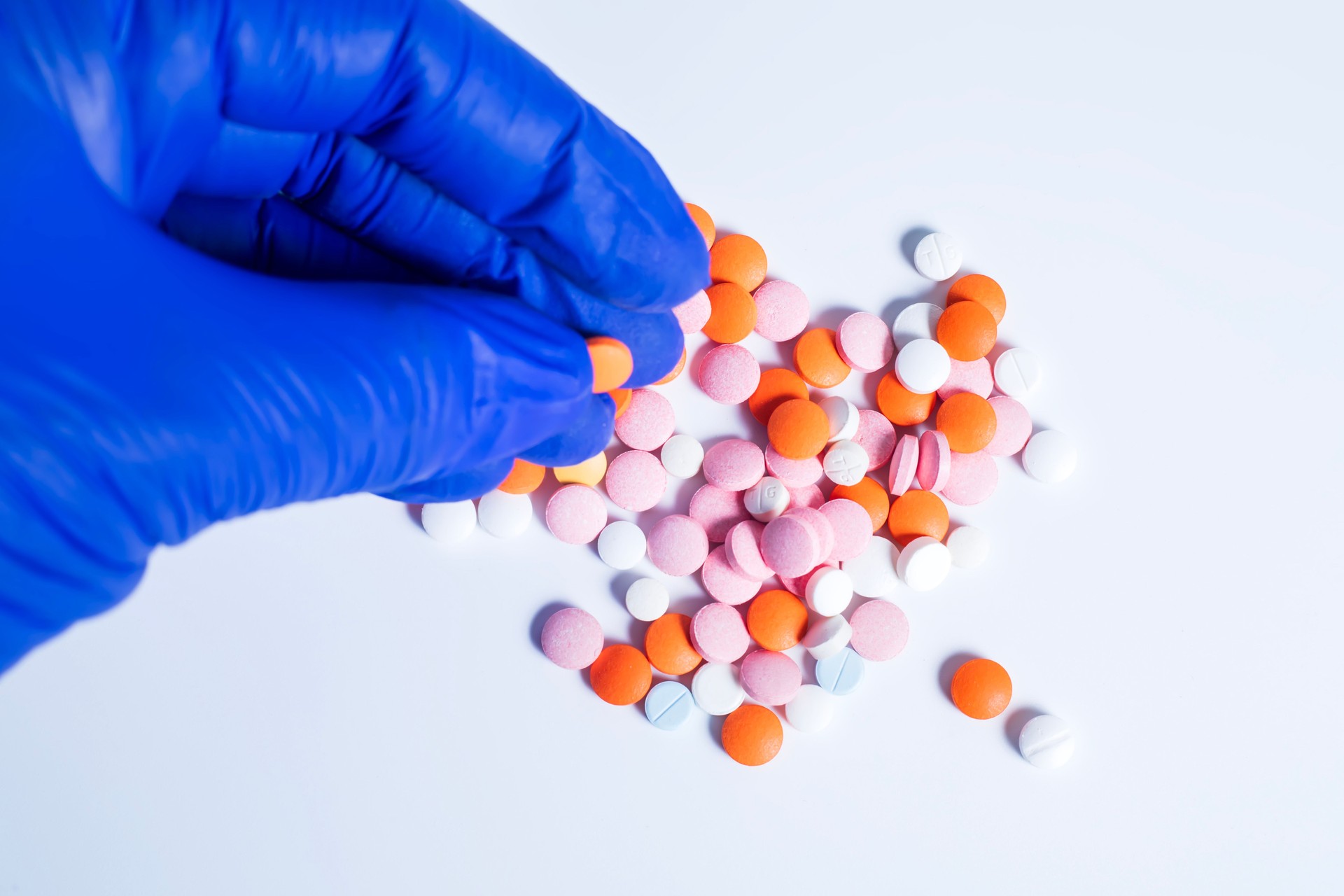 Doctor's hand in blue gloves holding orange color pill among multicolored pills lying on white table background. Top view